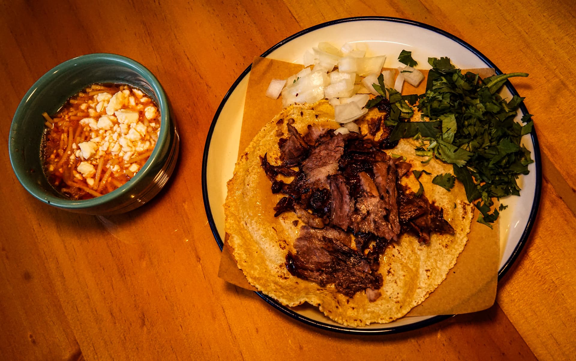 Taco with shredded meat, onion and cilantro beside a bowl of consommé
